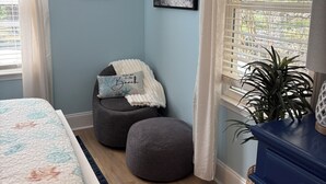 Ein Schlafzimmer mit einem Bett, einem grauen Hocker, einer blauen Kommode, einem Fenster mit Jalousien und einem gerahmten Bild an der Wand.