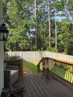 Eine Holzterrasse mit einem Korbstuhl und einem kleinen Tisch, mit Blick auf einen von einer Hecke umgebenen Garten mit Bäumen.