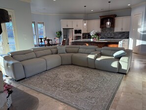 A spacious living room with a large L-shaped sectional sofa, a patterned rug, and a kitchen area with white cabinets and a stone backsplash.