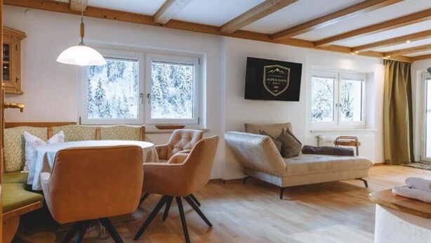 Ein gemütliches Zimmer mit Holzbalkendecke, einer Couch, einem Sessel und einem Tisch. Fenster bieten Blick auf schneebedeckte Bäume.
