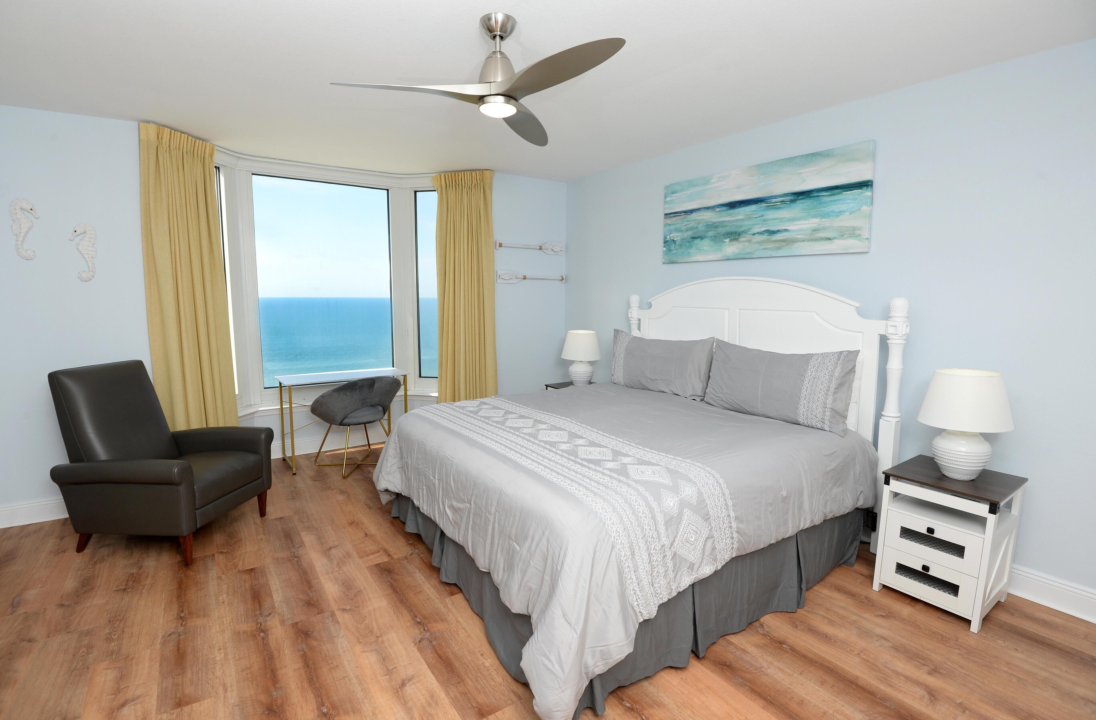 A bedroom with a bed, a chair, a nightstand, a ceiling fan, and a window with curtains.