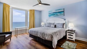Slaapkamer met een bed, nachtkastjes, een plafondventilator, een raam met gordijnen en uitzicht op de oceaan.