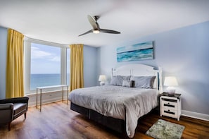 Ein Schlafzimmer mit Bett, Nachttischen, Deckenventilator, einem Fenster mit Vorhängen und Meerblick.