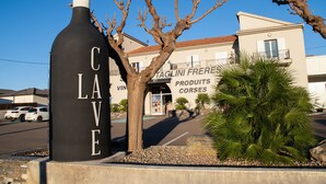 A large black bottle-shaped sign with the word 'CAVE' on it, a tree, and a building with 'TAGLINI FRERE PRODUCTS CORSES' signage.