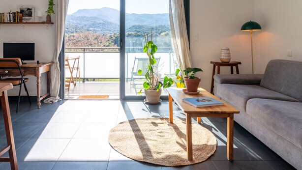A modern living room with a sofa, a wooden desk, a television, and a balcony with a view of mountains.