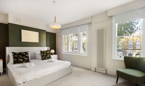 A modern bedroom with a large bed, a green armchair, and a view of buildings through large windows. - 10 Mins Walk to The Oval The Kennington Retreat (London)