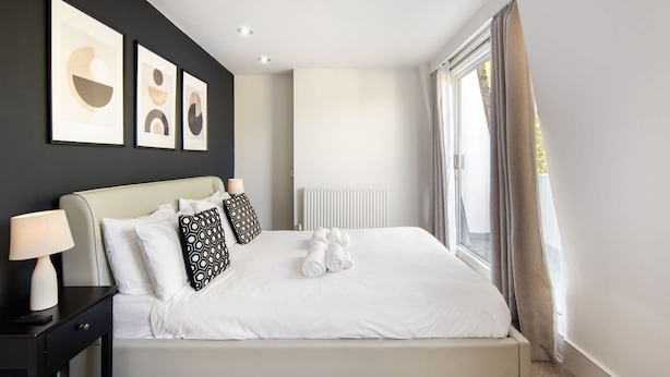 A neatly made bed with white linens and two patterned pillows, a bedside lamp, and a framed abstract artwork on the wall. - 10 Mins Walk to The Oval The Kennington Retreat (London)