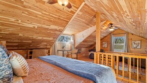 Um quarto de sótão aconchegante com teto de madeira, cama com roupa de cama estampada, escrivaninha e janela com cortinas.
