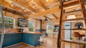 Interior de cabana de madeira com cozinha, armários azul-turquesa, espaço para café e área de jantar com escada e cadeira.