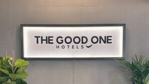 A reception area with a framed sign reading "THE GOOD ONE HOTELS" above a dark leather sofa. There are two potted plants on either side of the sofa. - THE GOOD ONE Hotel  (Landshut)