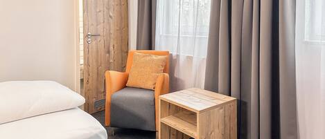 Une pièce comprenant un lit, une chaise en bois, une petite table en bois et une fenêtre avec des rideaux.
