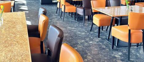 Un interno ristorante moderno con travi in legno a soffitto, lampade a sospensione rotonde e un mix di sedie in pelle color arancione e marrone attorno a tavoli quadrati.