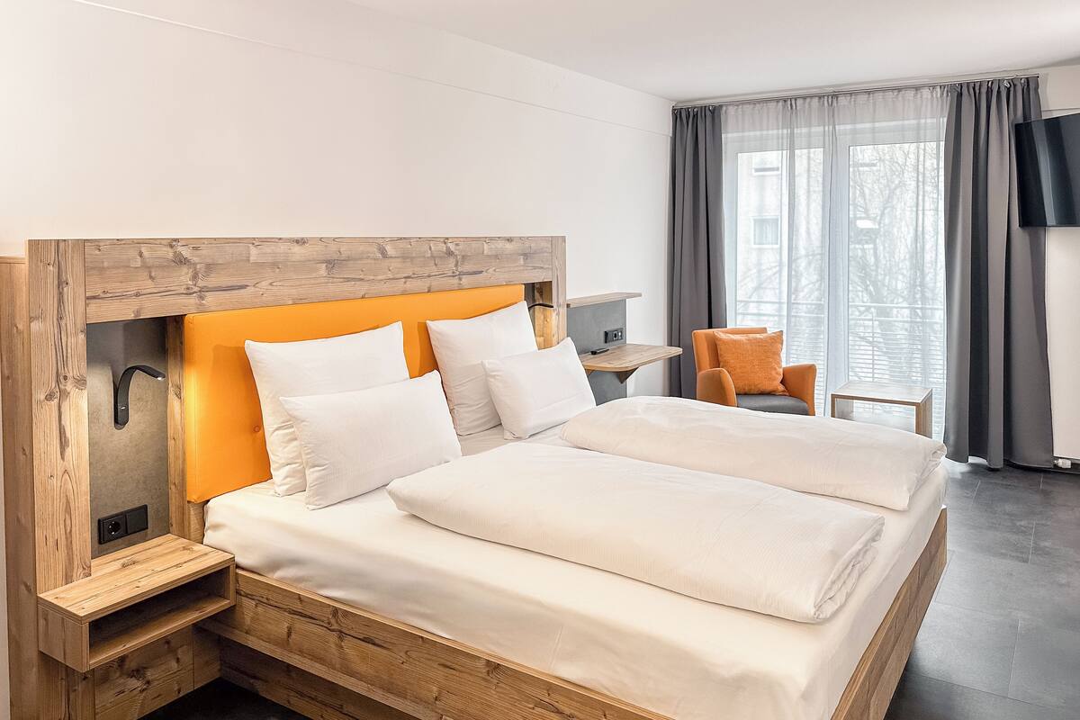 A modern hotel room with a large bed, wooden headboard, and a seating area with a chair and table.