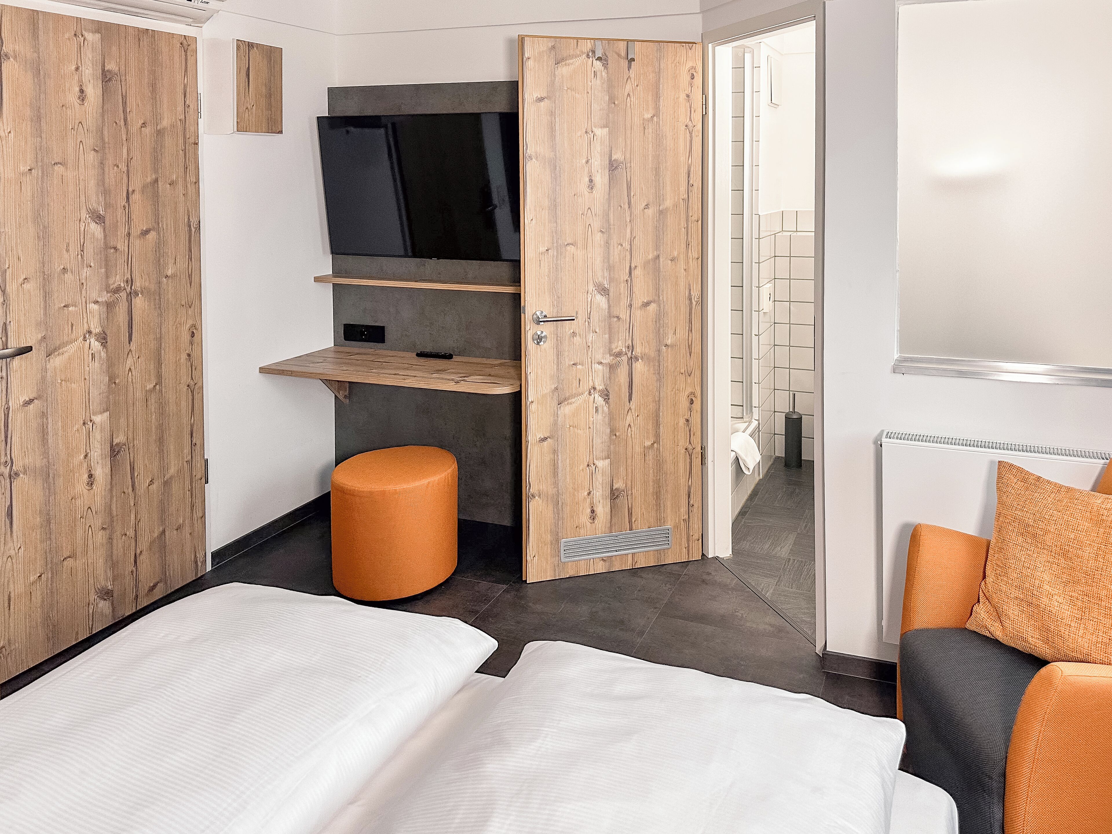 A modern hotel room with two beds, a wooden wardrobe, a small orange stool, and a TV mounted on the wall.