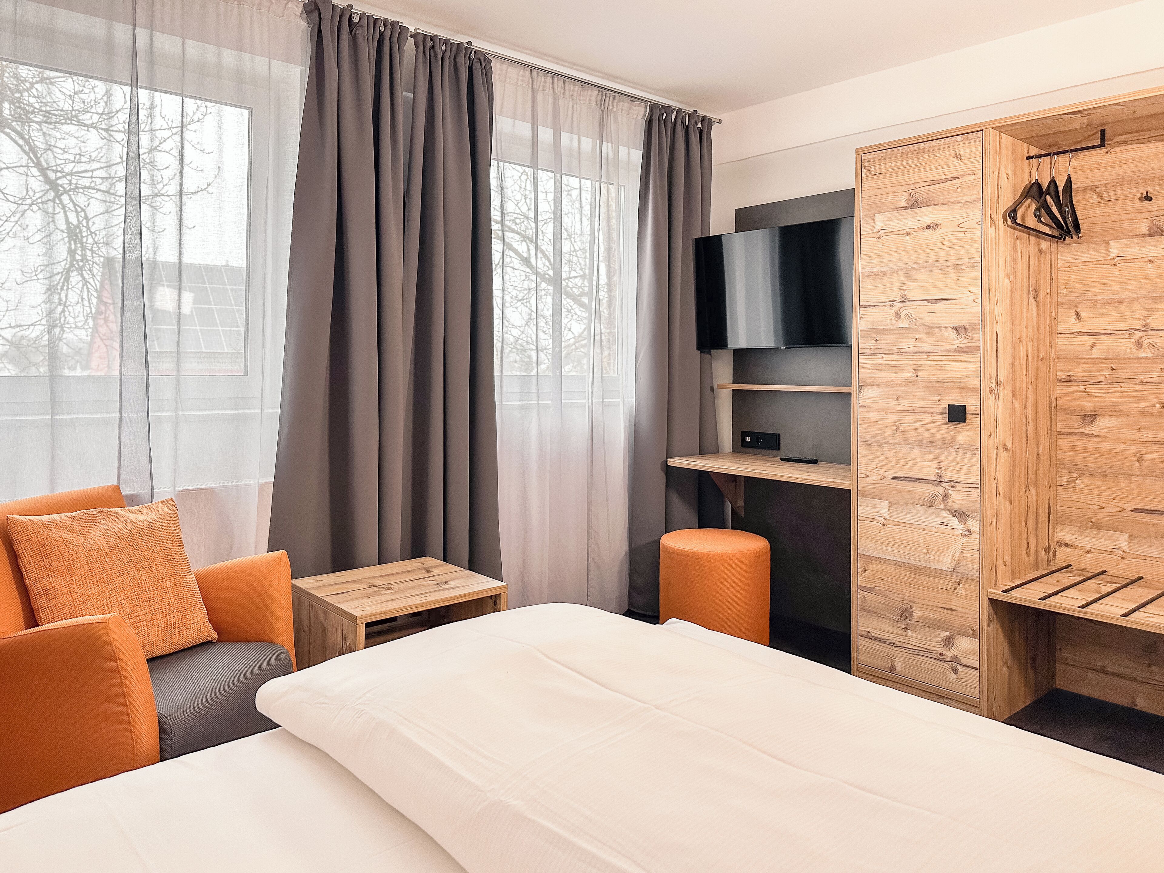 A modern hotel room with a bed, a seating area with orange cushions, a wooden wardrobe, a TV, and a small table.