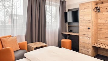 Una moderna camera d'hotel con un letto, una zona salotto con cuscini arancioni, un armadio in legno, una TV e un tavolino.