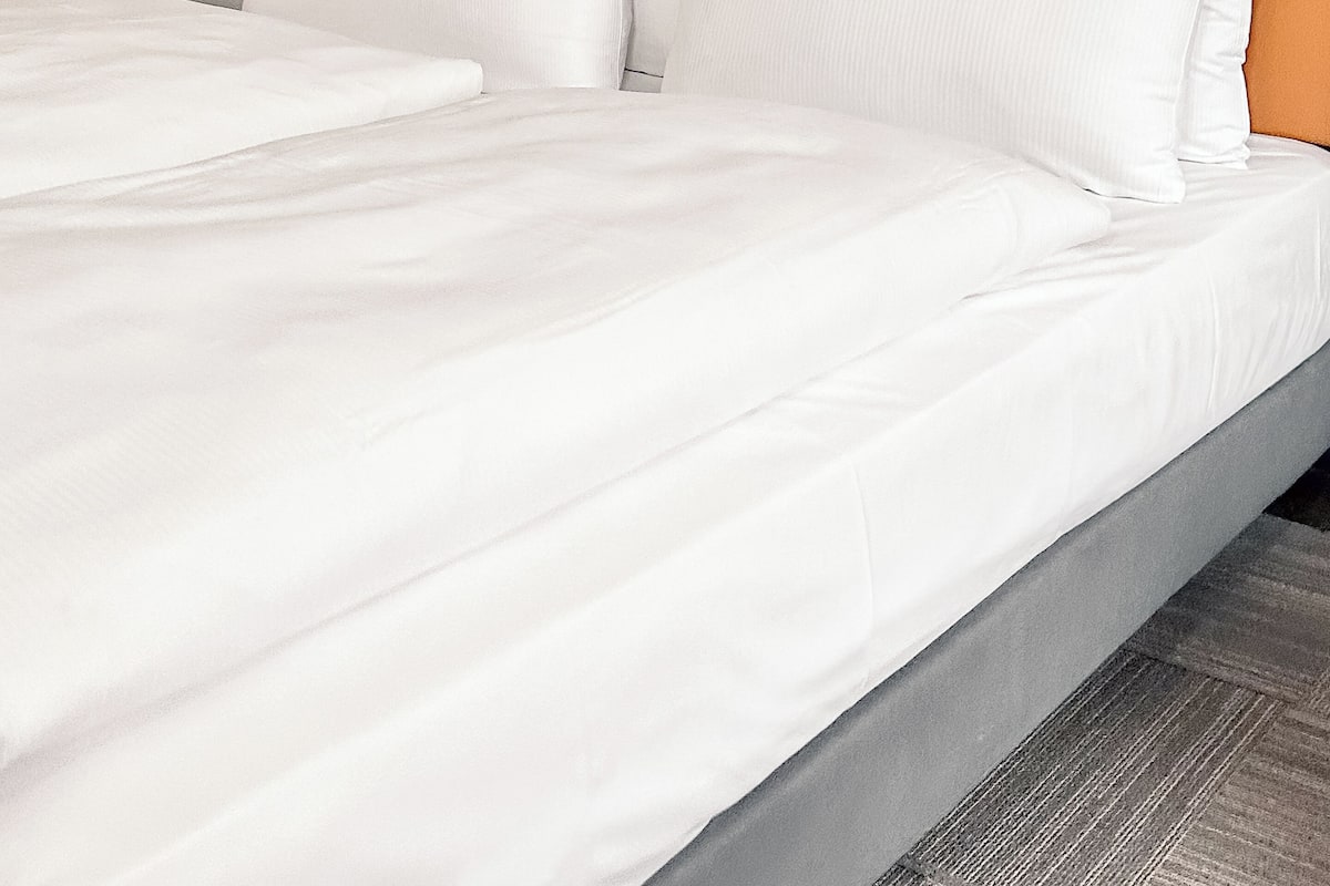 A neatly made bed with white linens and pillows, a wooden headboard, and a framed artwork above it.