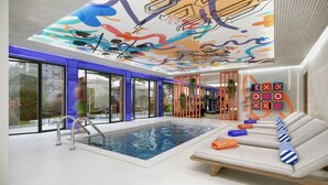 A modern indoor pool area with a glass wall, a lounge with a sofa, and a ceiling with colorful artwork. - Symone Poitiers (Poitiers)