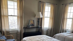 Un dormitorio con una cama, una silla, una mesita de noche con lámpara y una ventana con cortinas.