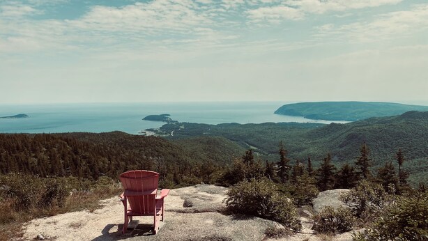 Een rode Adirondack-stoel op een rotsachtige richel met uitzicht op een kustlandschap met dichte bossen en verre eilanden.