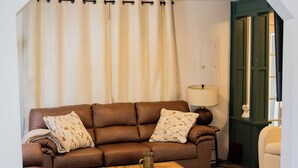 Een woonkamer met een bruine bank, een houten salontafel en een kamerbreed tapijt. Aan de muur hangen ingelijste kaarten en op een bijzettafeltje staat een lamp.