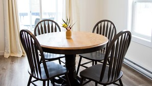 Een eetruimte met een ronde houten tafel, omringd door vier stoelen, een vaas met bloemen op de tafel en grote ramen met gordijnen.