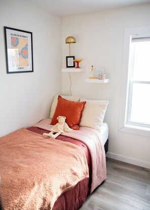 A bedroom with a bed, pillows, a teddy bear, a framed picture, a lamp, and a window.
