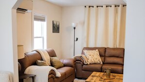 Een woonkamer met een bruine leren bank, een houten salontafel en een kamerbreed tapijt. Er hangen ingelijste kaarten aan de muur en er staat een vloerlamp.
