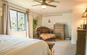 Un dormitorio con una cama, una silla, un aparador, un ventilador de techo y una ventana con cortinas.