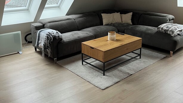 Moderni loft-tyylinen asunto, jossa on suuri harmaa kulmasohva, puinen sohvapöytä ja näkymä kaupunkimaisemaan suurten kattolasien kautta.