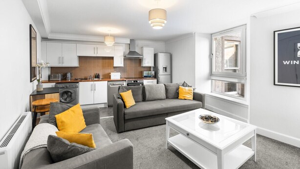 Eine moderne Wohnung mit Wohnzimmer, Küche und Essbereich. Sie verfügt über ein graues Sofa, einen weißen Couchtisch und ein großes Fenster, das für natürliches Licht sorgt.
