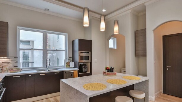 Une cuisine moderne avec des meubles de couleur sombre, un îlot en marbre, des luminaires suspendus et une vue sur la ville.