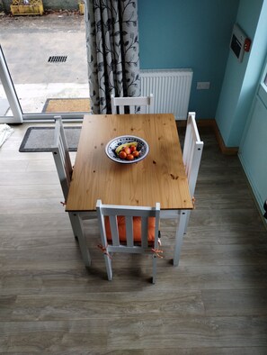 A dining area with a wooden table and white chairs, a bowl of fruit, and a view of the outside.