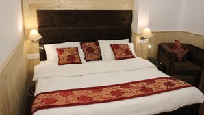 Un lit avec une tête de lit en bois, une literie blanche et des coussins à motifs floraux rouges. Il y a deux lampes de chevet et une petite table avec une chaise.