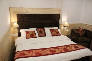 A bed with a wooden headboard, white bedding, and red floral pillows. There are two bedside lamps and a small table with a chair.
