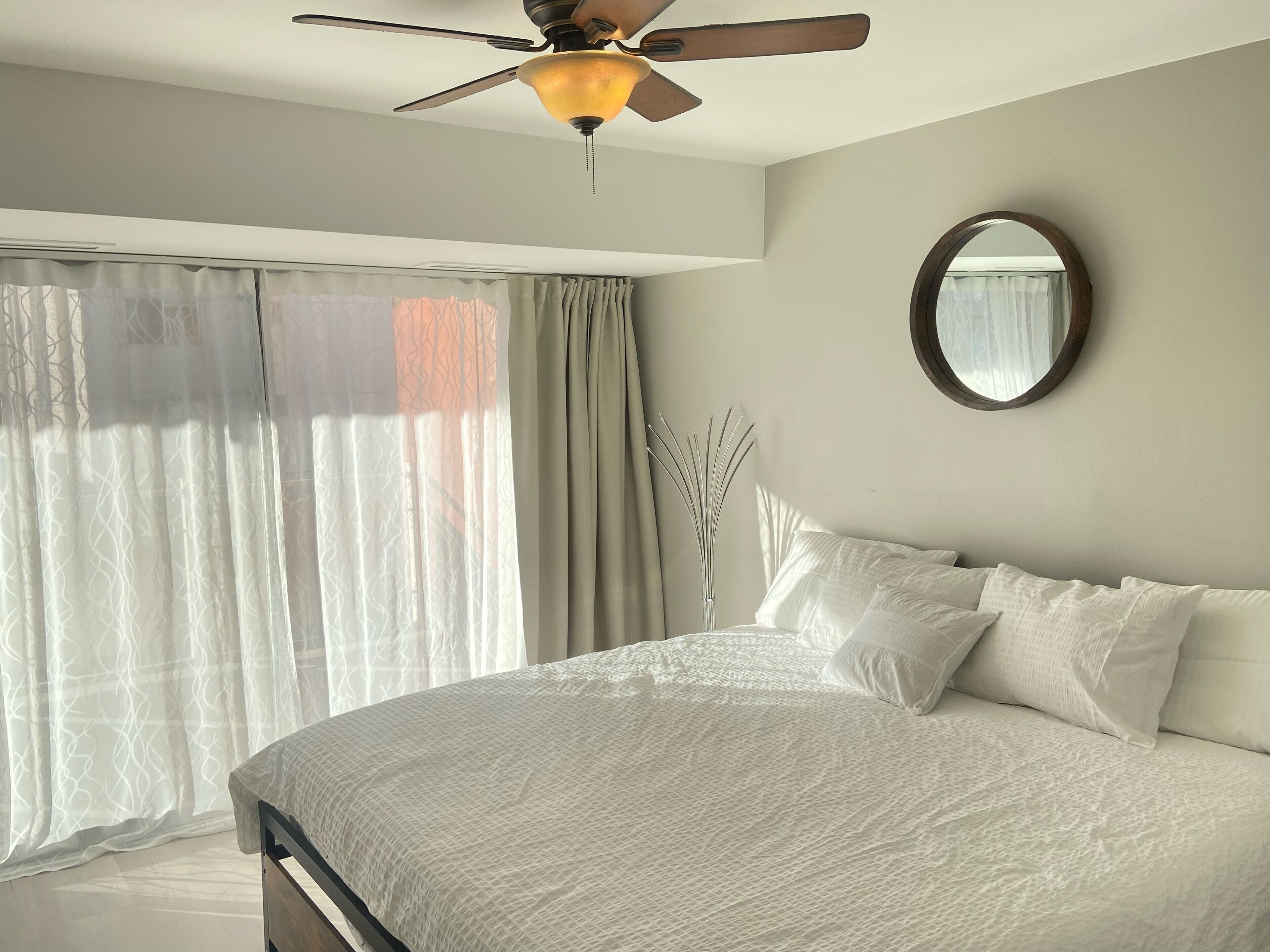 Quarto com cama, ventoinha no teto, janela com cortinas e espelho redondo na parede.