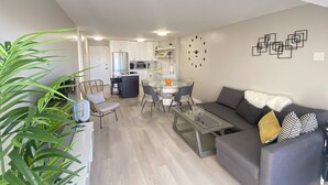 A modern living room with a glass coffee table, grey sofa, and a dining area with a white table and chairs. - Lovely 1-bedroom condo near shops & dining! (Denver)