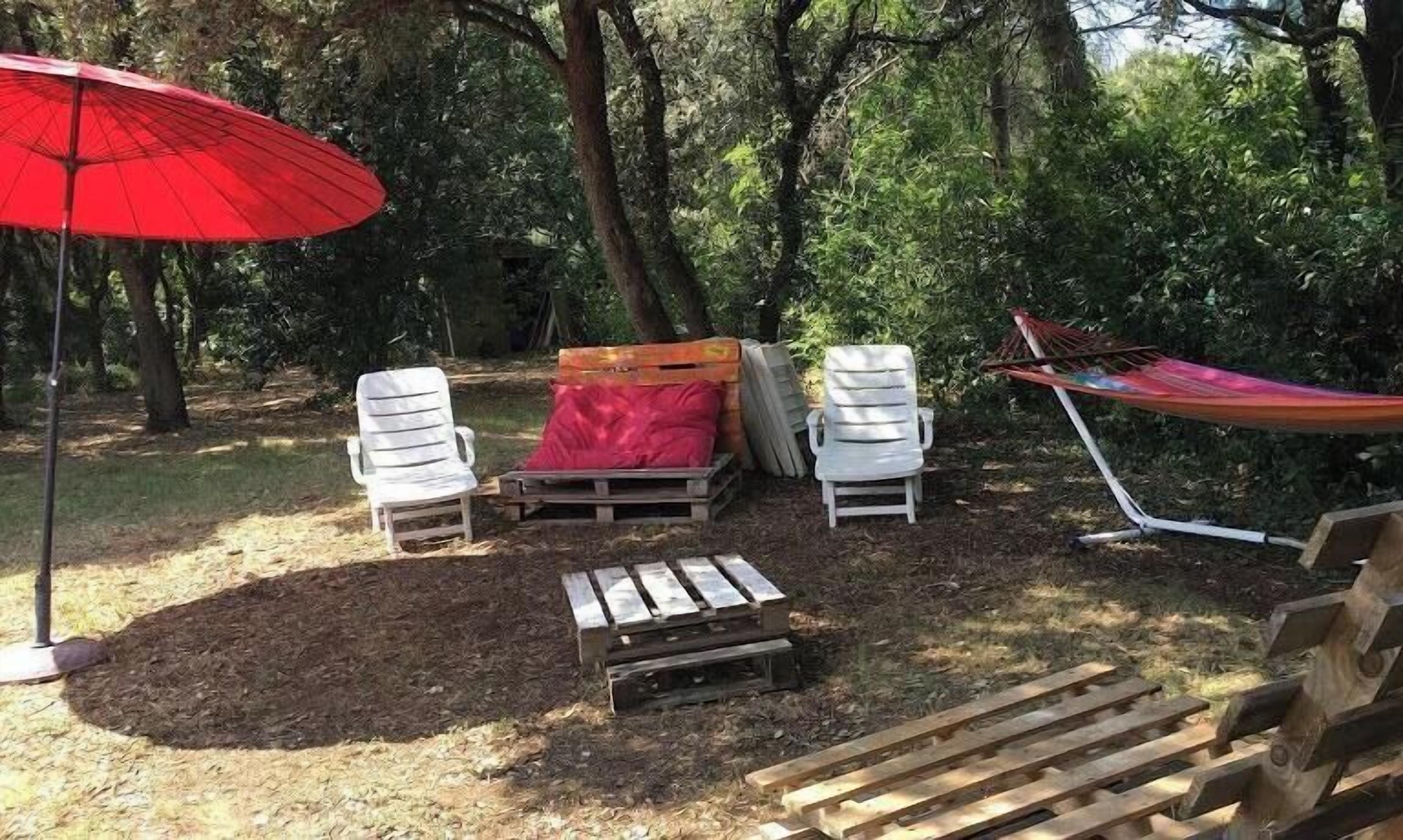 Un jardin avec un parasol rouge, deux chaises blanches, un banc en bois et un hamac.