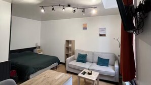 A compact room with a bed, a sofa, a small table, a TV mounted on the wall, and a ceiling light fixture. - L’Hermione – city center & thermal baths (Rochefort)