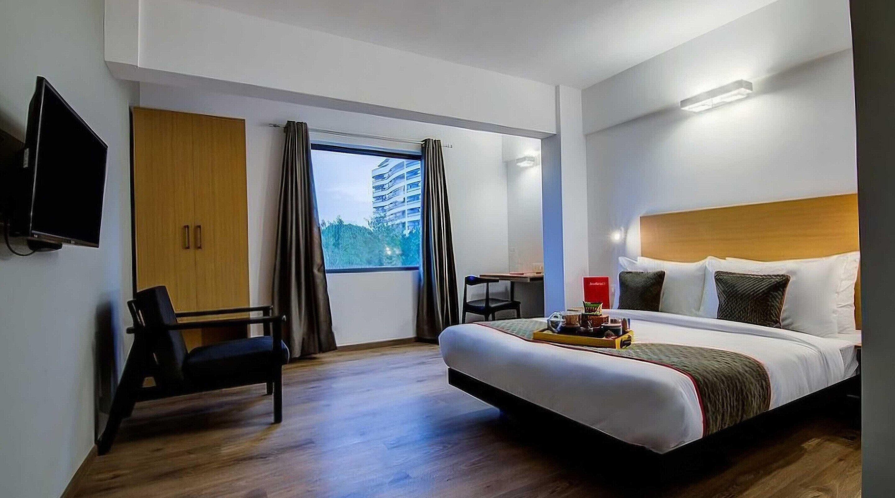Habitación de hotel con cama, escritorio, silla, televisor y ventana con cortinas.