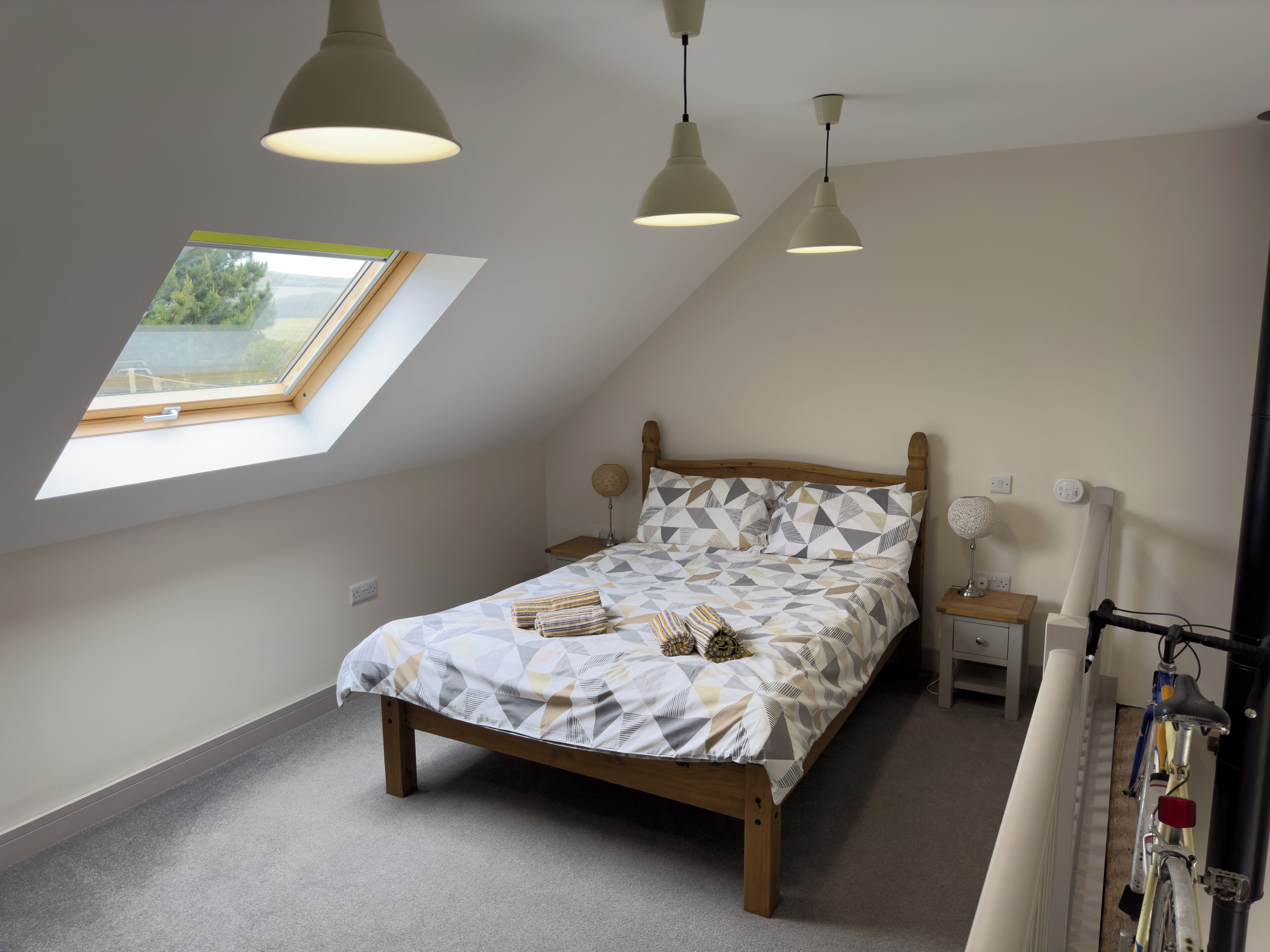 A bedroom with a sloped ceiling, a skylight, a bed with patterned bedding, a bedside table with a lamp, and a bicycle rack.