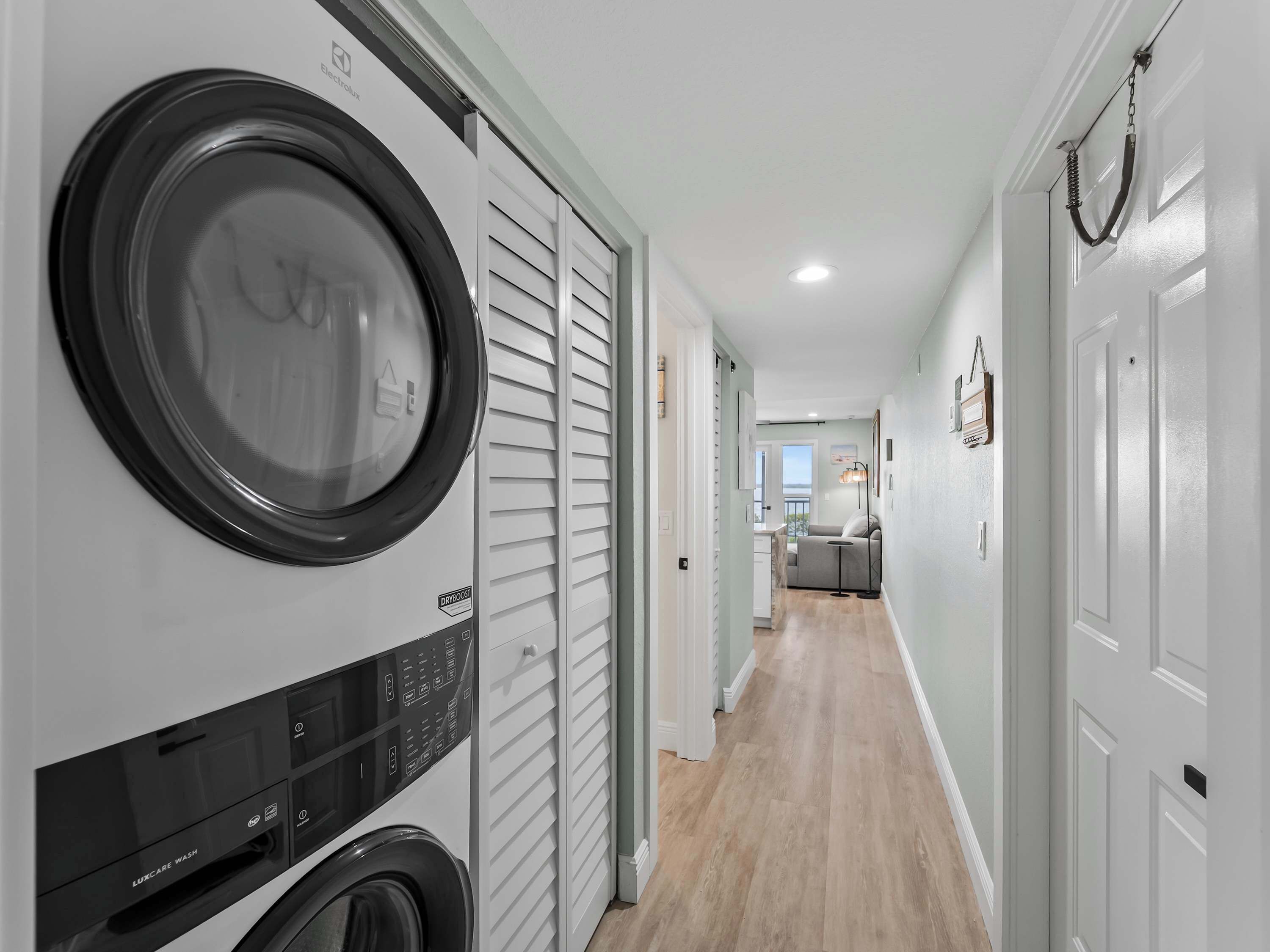 A laundry room with a front-loading washer and dryer, a hallway leading to a kitchen, and a door with a hanging towel rack.