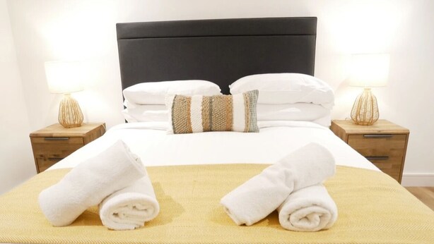 A neatly made bed with white linens and two rolled towels on a yellow bedspread. There are two bedside tables with lamps, one on each side of the bed. - Scandic House Apartments - Long Term Rental Available (Ulverston)