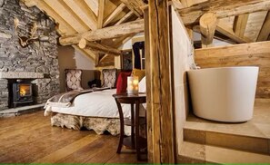 A cozy attic bedroom with a stone fireplace, wooden beams, a bed with a white comforter, and a large white chair. - Presidential Suite (Sainte-Foy-Tarentaise)