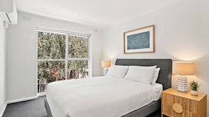 Una habitación de hotel moderna con una cama grande, una mesita de noche con lámpara, una obra de arte enmarcada y vistas a zonas verdes a través de un ventanal amplio.