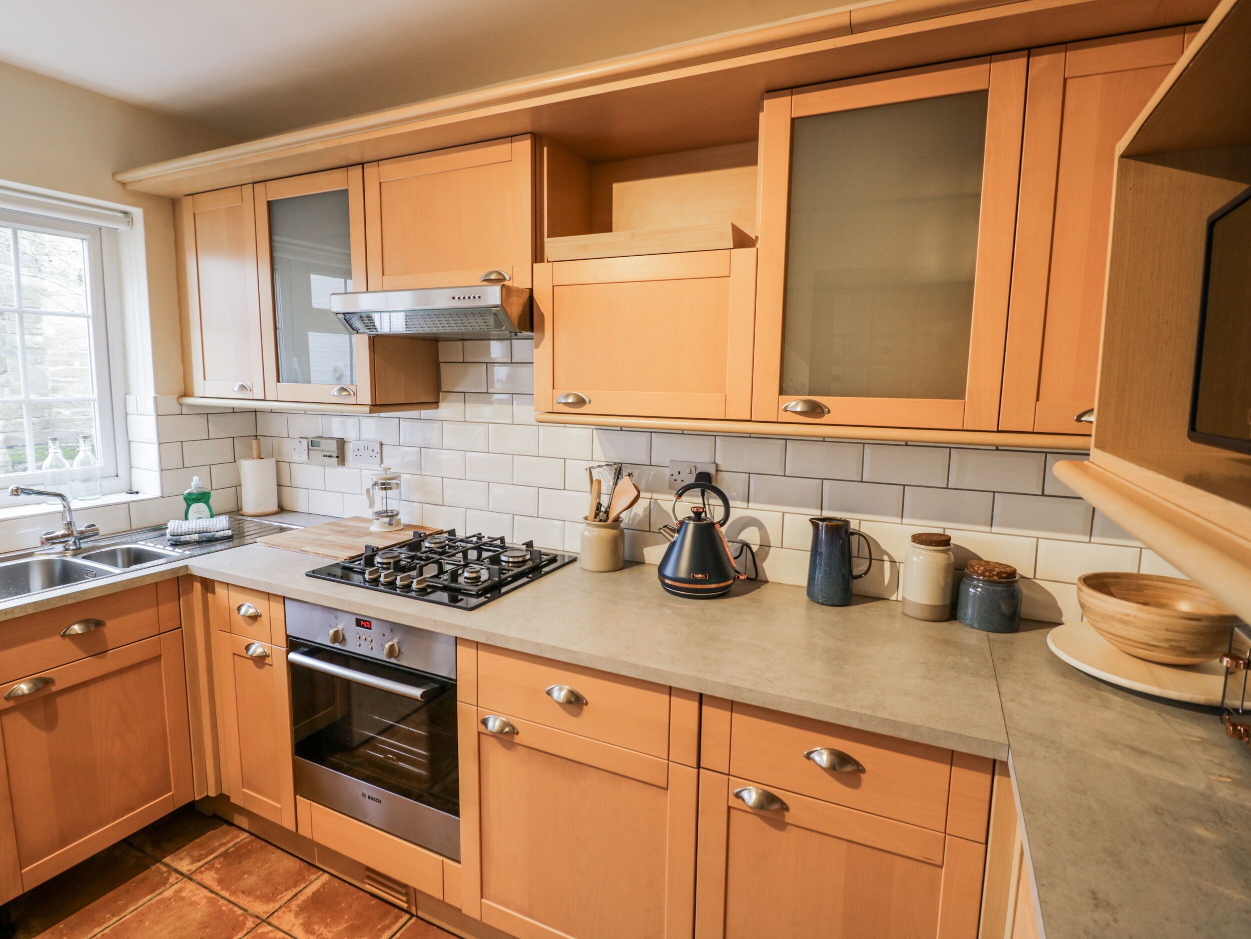 Une cuisine moderne avec des meubles en bois, un four Bosch, une cuisinière à gaz et une fenêtre qui laisse entrer la lumière naturelle.