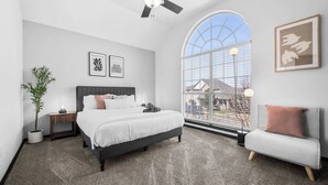 A bedroom with a bed, a nightstand, a potted plant, a gray sofa, and a large window with a view of buildings.