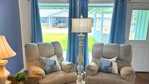 Quarto com duas poltronas bege, abajur, mesa pequena com luminária e vista para uma casa através de cortinas azuis.