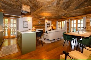Ein rustikaler Innenraum mit Holzbalkendecke, einer Küchenzeile mit Kochinsel, einem Esstisch mit Stühlen und einer Couch.