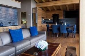 Ein modernes Wohnzimmer mit einem grauen Sofa, blauen Kissen und einem Couchtisch mit Blumen. Im Hintergrund ist ein Essbereich mit blauen Stühlen zu sehen.